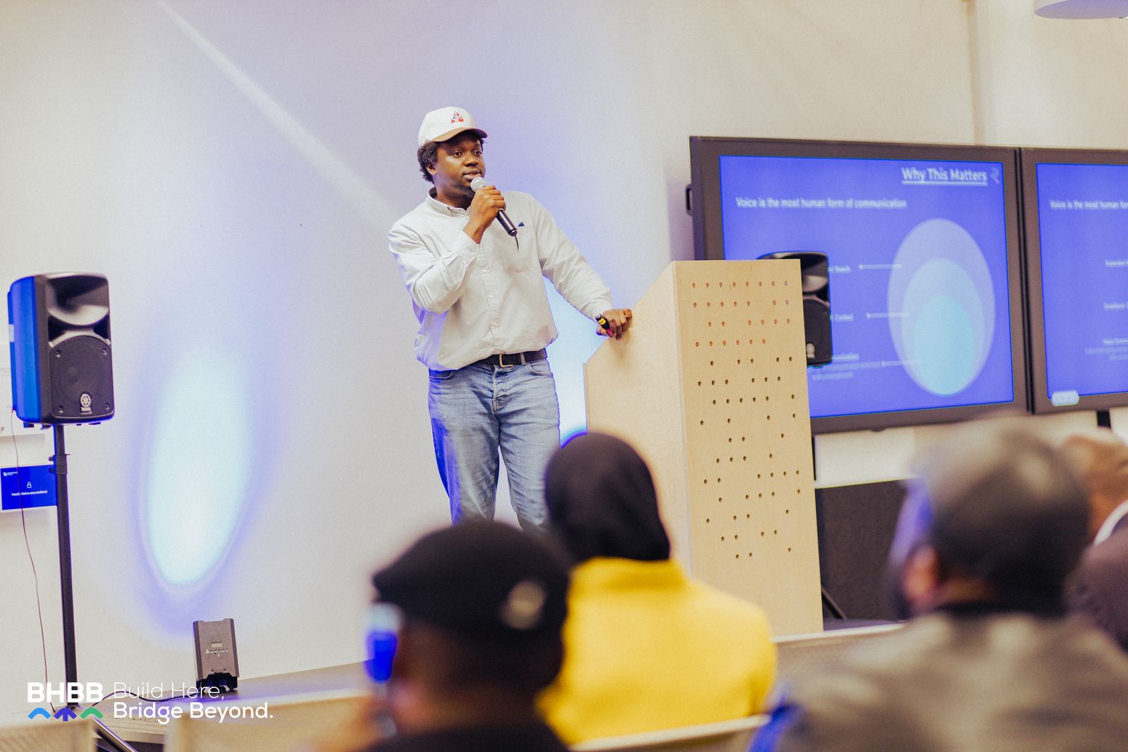 Ikechukwu on stage with a microphone beside a podium, presenting a slide titled 'Why This Matters' — 'Voice is the most human form of communication' — to a seated audience